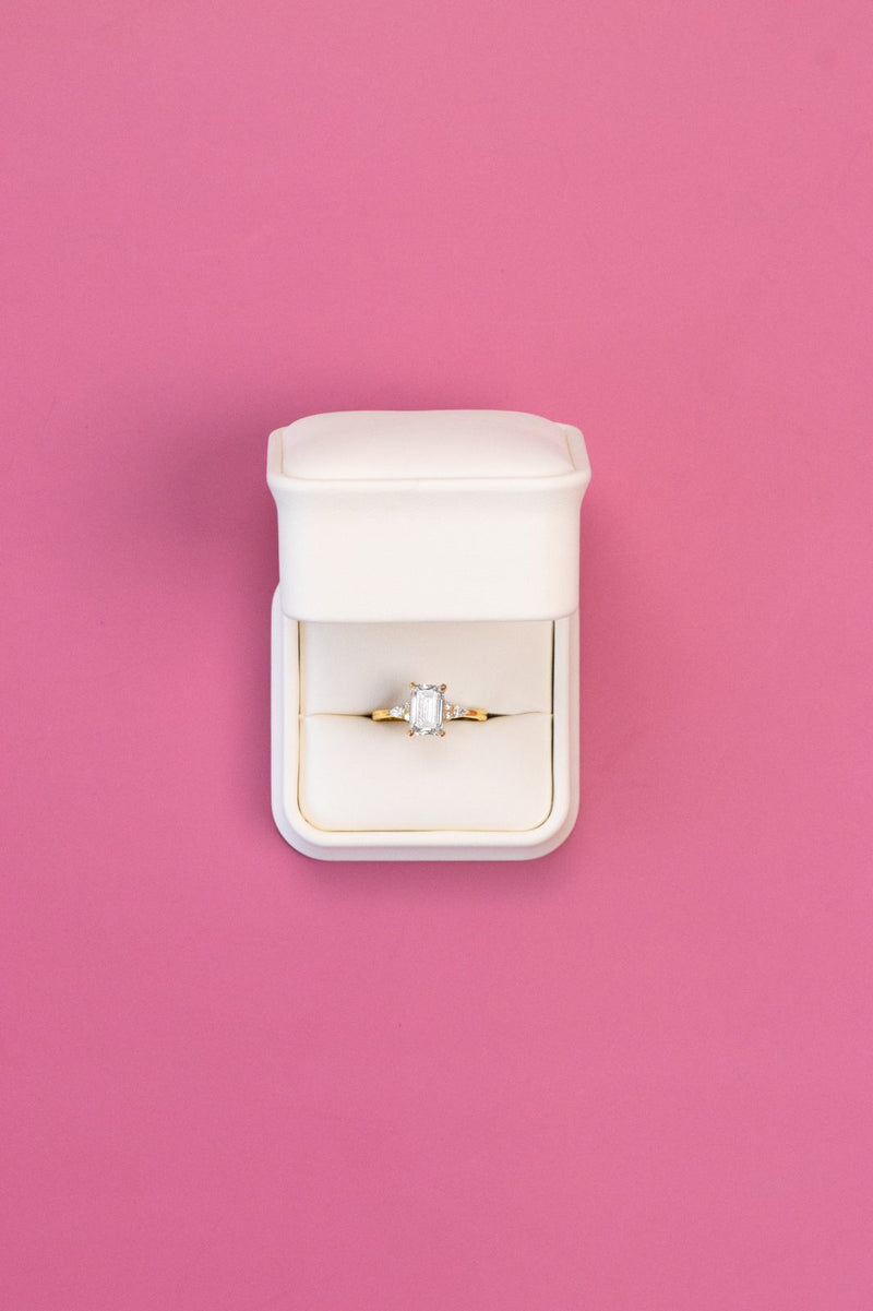 Emerald-cut ring with side stones in a white box on a pink backdrop