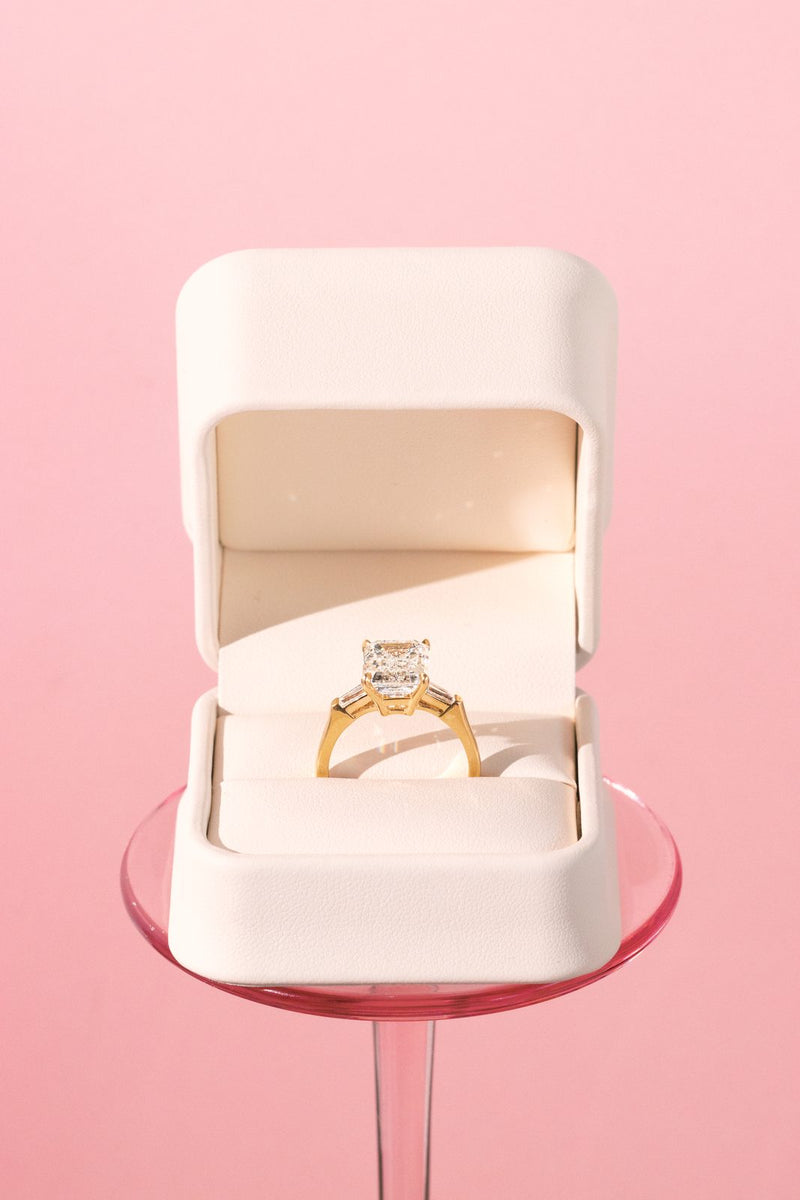 Emerald-cut ring in a white box raised on a glass stand against pink